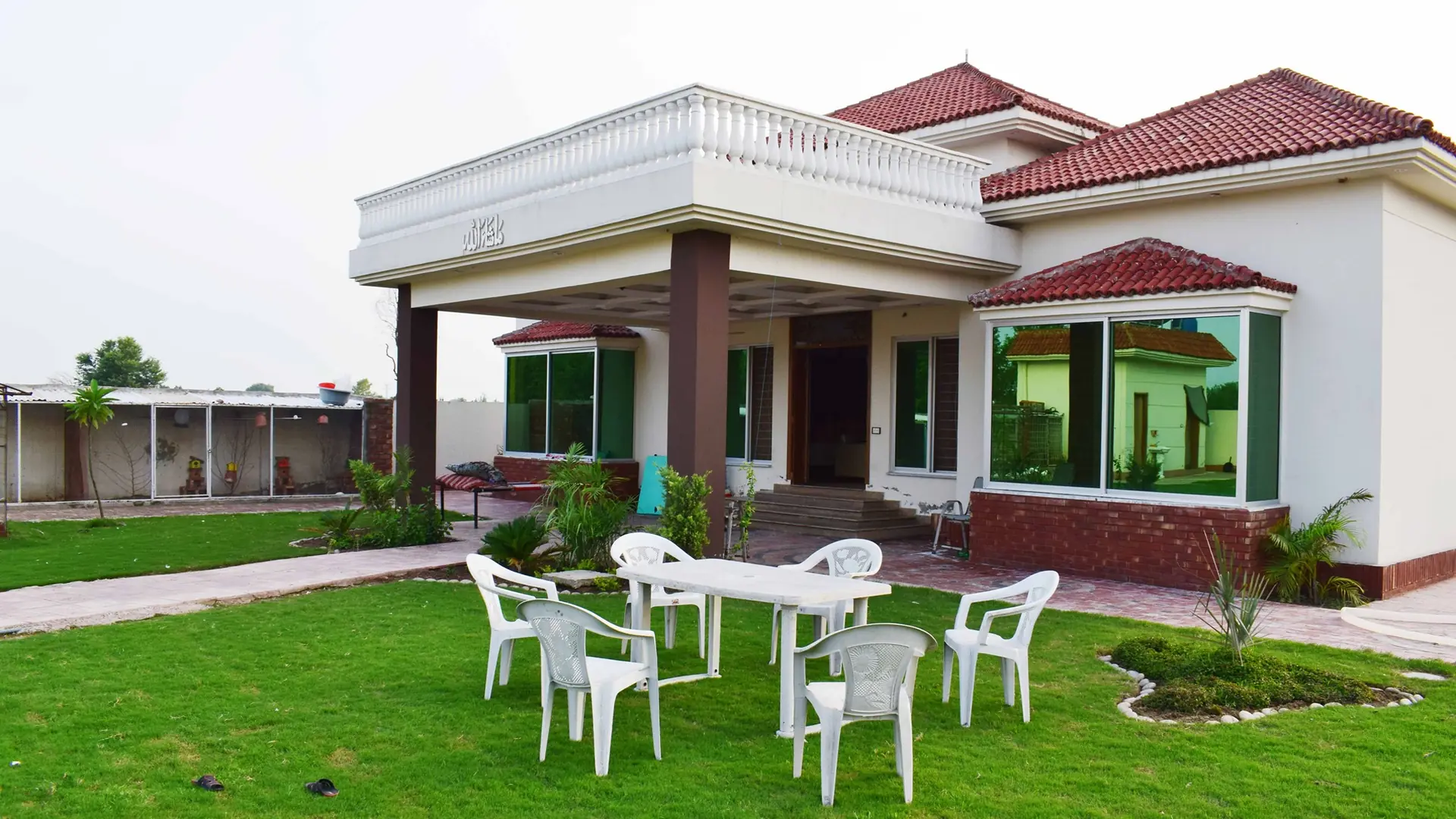 Moments Farmhouse on Barki Road, Lahore: the white villa with red tiled roofs behind a green lawn with garden chairs